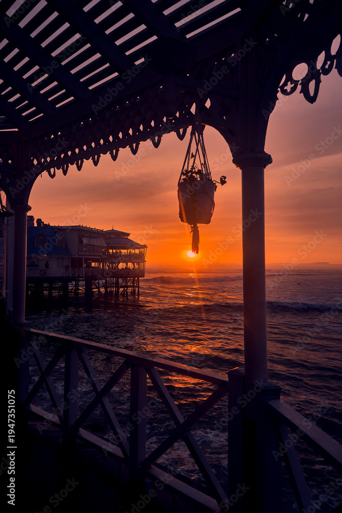 Fototapeta premium Golden sunset light paints the Pacific Ocean as waves roll beneath the iconic La Rosa Nautica restaurant on the Miraflores coast in Lima, Peru. The tranquil seaside atmosphere evokes an active coastal