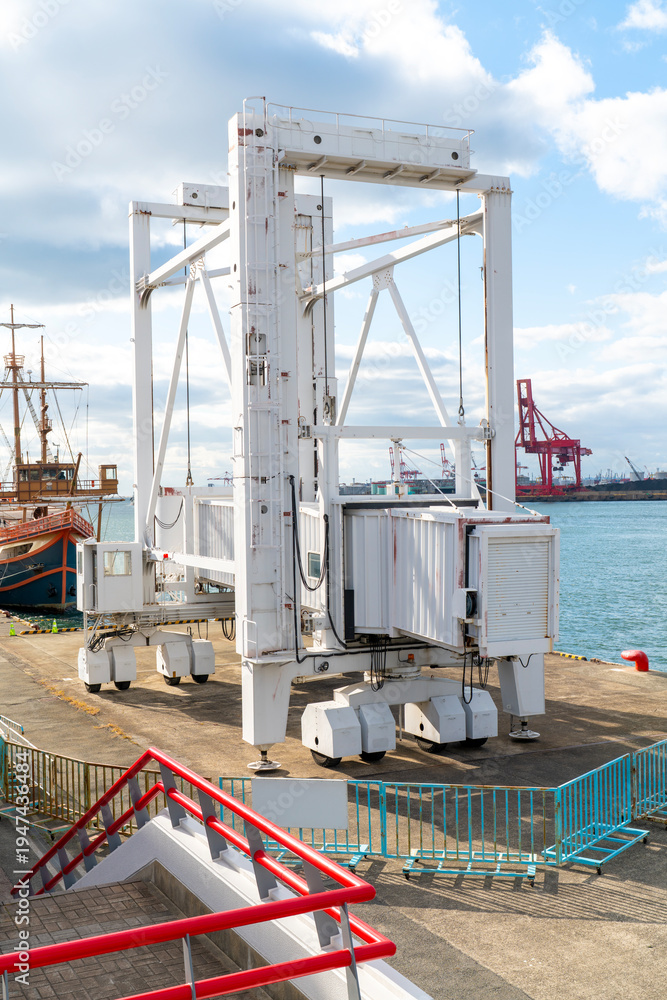 Fototapeta premium Large white mobile passenger gangway on wheels at a busy harbor. Professional boarding bridge for ships located on a dock under a bright sky with industrial port equipment in the background.