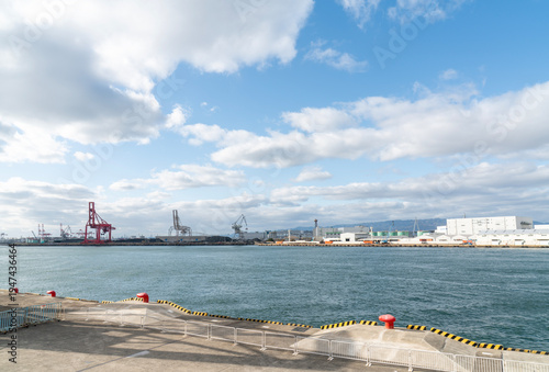 Wide angle view of a commercial industrial port featuring shipping cranes and warehouses across the sea. Concrete dock foreground with red bollards and barriers under a blue sky with clouds.