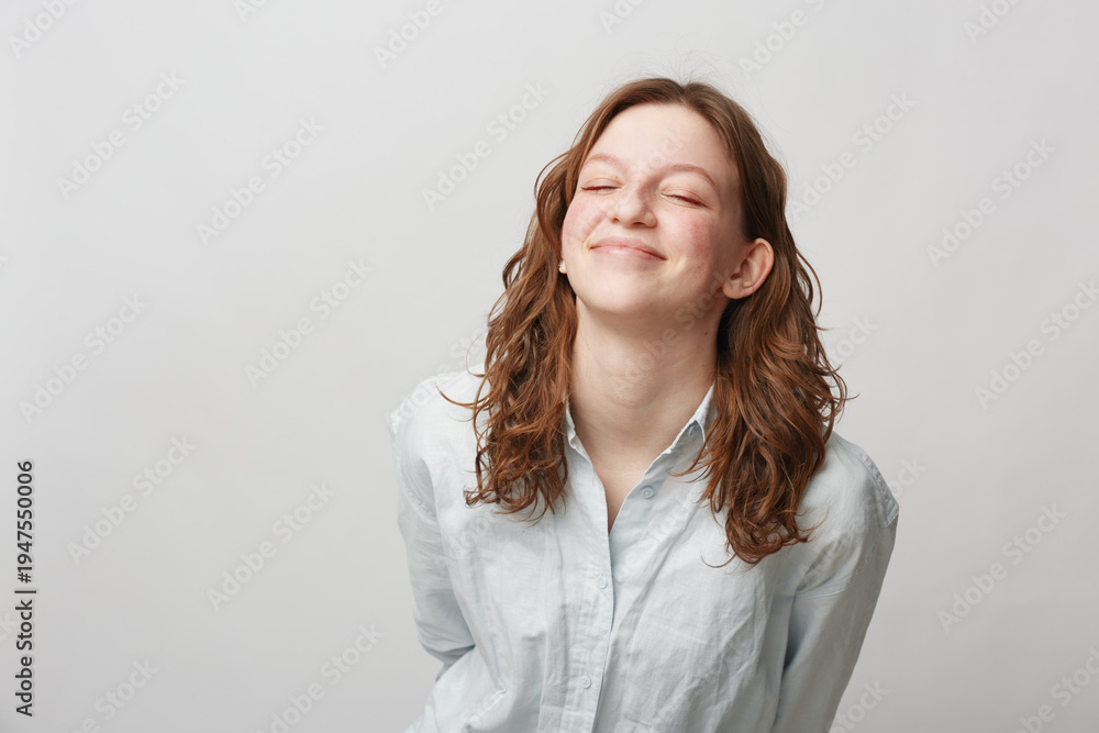Fototapeta premium Young woman with curly hair smiles with closed eyes wearing white shirt on light background. Concept of happiness, relaxation, and positive emotions in casual lifestyle.