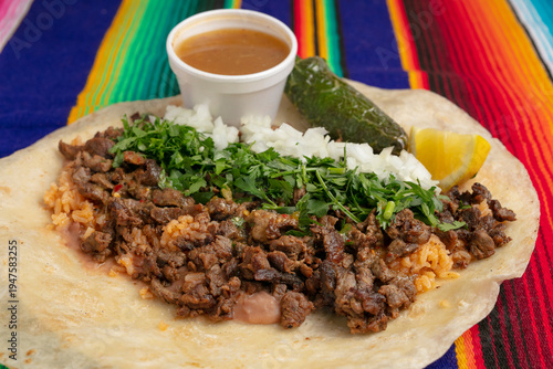 Close up shot of an open carne asada burrito with cilantro, onion, lime, salsa, rice and jalapeño on a flour tortilla and traditional Mexican tablecloth 