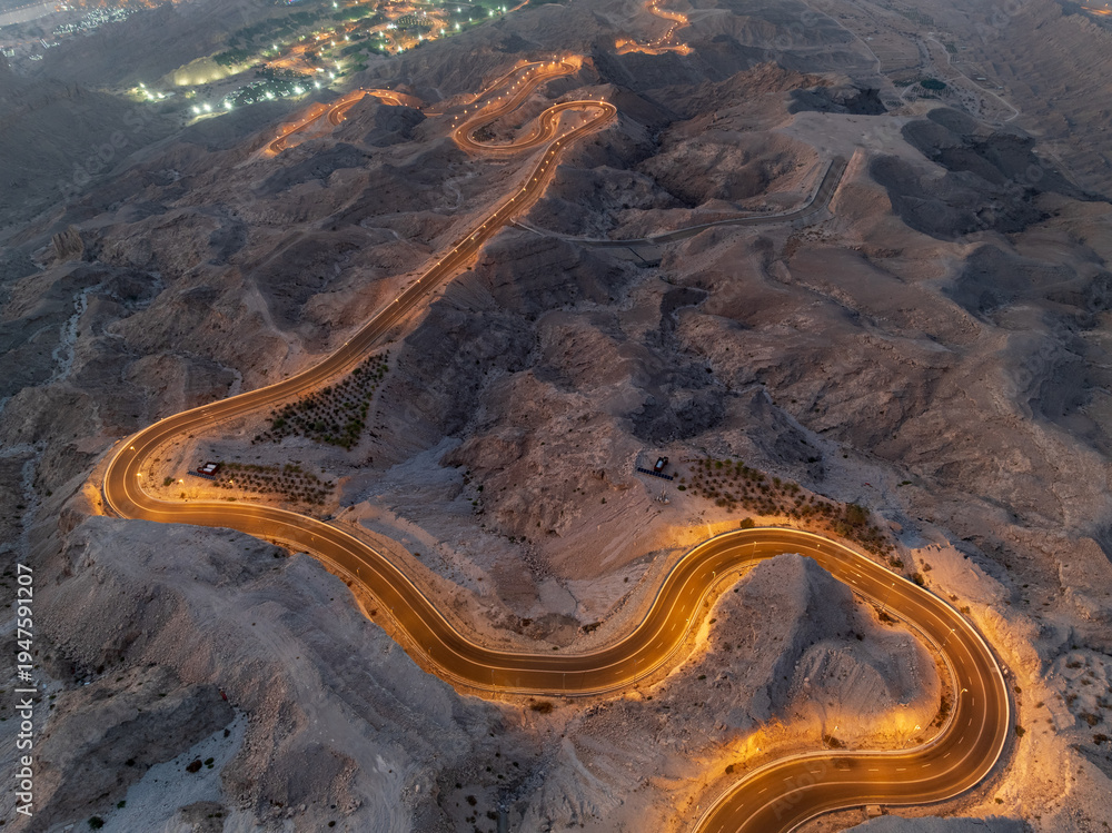 Naklejka premium An aerial view of a winding road through a desert landscape at dusk with city lights in the distance