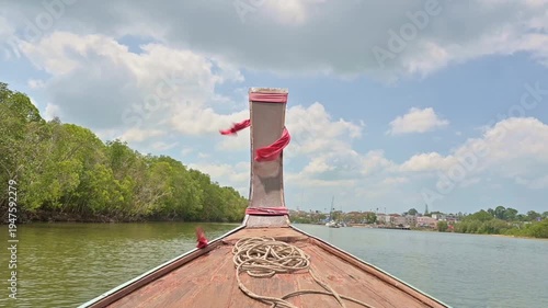 Front view of wooden long tail boat cruising in the sea along coastal heading to Krabi Town. Travel destination surrounded by green vegetation in natural canal under cloudy blue sky in Thailand. 