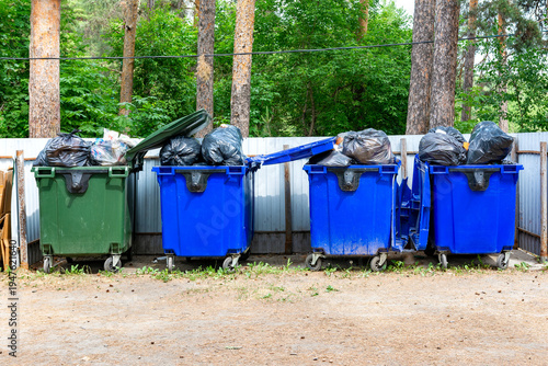 A row of overflowing dumpsters stands by the fence in a public park, causing environmental pollution.	