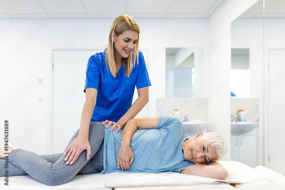 Fototapeta premium Professional female therapist performing manual hip therapy on a senior woman in a bright clinic. Specialist assisting an elderly patient with lower back and pelvic stability exercises.