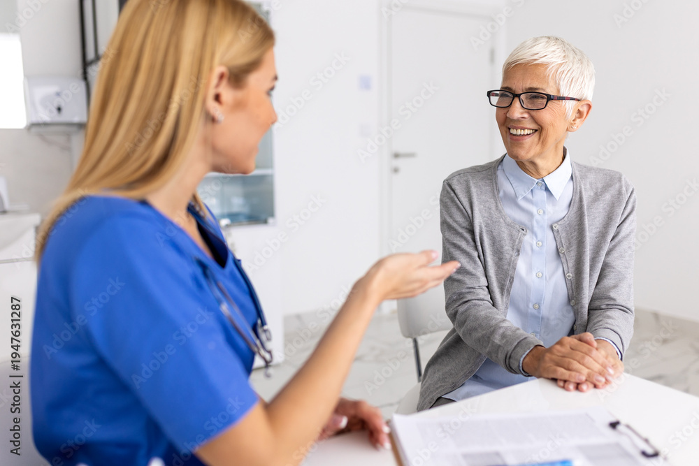 Fototapeta premium Happy senior woman smiling during consultation with female doctor in blue scrubs friendly medical professional providing geriatric care and emotional support in bright hospital office.