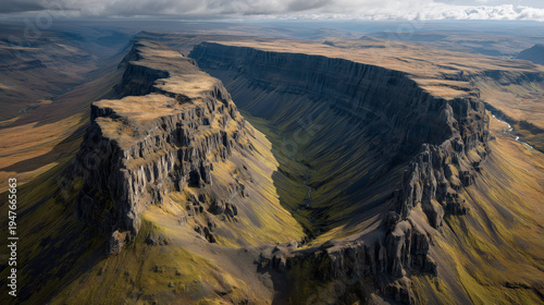 Dramatic aerial view of a vast, rugged canyon with steep cliffs and sparse vegetation under a cloudy sky.
