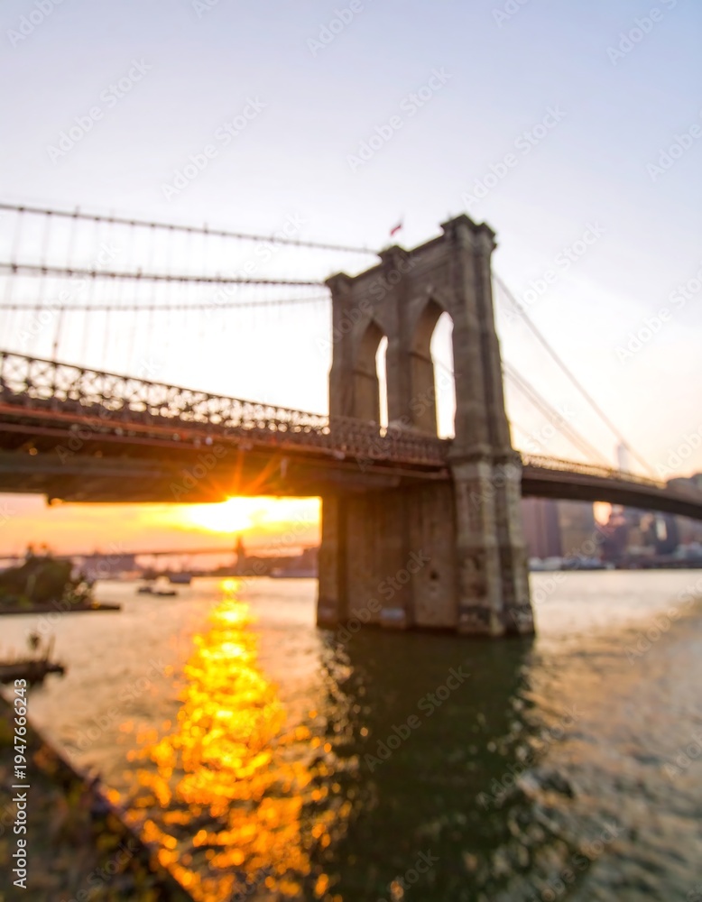 Fototapeta premium Blurry photograph of a large suspension bridge in New York City at sunset, golden light reflecting off the water below