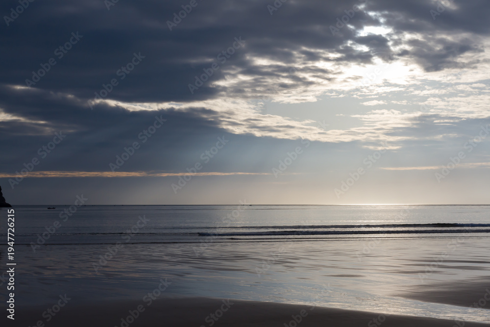 Obraz premium Dramatic sunlight breaking through dark clouds over the ocean and beach.