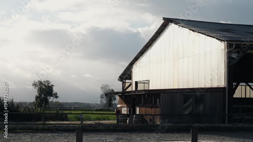 Wallpaper Mural Wide exterior shot of a large wooden and metal horse stable building. A professional riding arena with wooden fences is visible in the foreground against a rural landscape. Torontodigital.ca