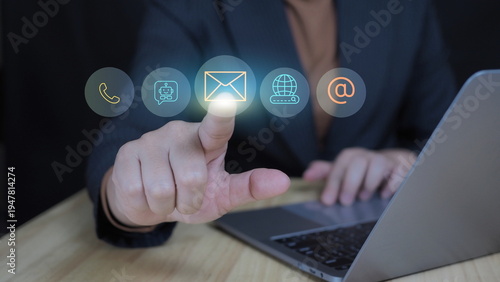 Businesswoman touching glowing email icon on virtual contact interface with phone, chatbot and web symbols while using laptop. Concept of digital communication, customer support.