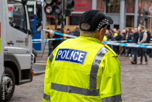 Quadro em tela Police officer in a high-visibility jacket at a cordoned city street scene with an emergency vehicle and a crowd