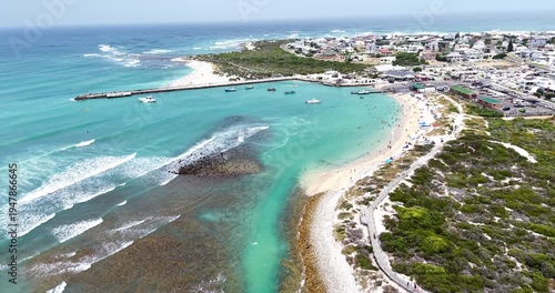 Drone aerial view of the harbour and beach at Struisbaai, showing turquoise water, small fishing boats, and beachgoers enjoying the sandy shoreline near Cape Agulhas on South Africa’s southern coast.
