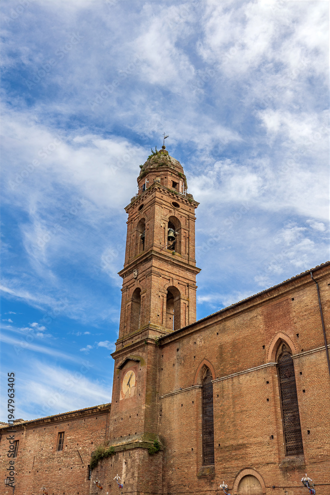 Fototapeta premium The Church of San Niccolo al Carmine shows its red brick structure under a blue sky. The tall bell tower features a clock. This site is part of Tuscany's architectural heritage in Siena.