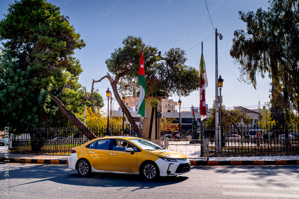 Obraz premium Taxi drives through a roundabout in the Jordanian capital with Jordanian flag on the street. Transport in Amman.