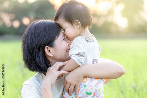 Asian mother holding her baby with a loving forehead touch. Warm family moment showing motherhood, bonding, affection, and happy parenting outdoors in natural light