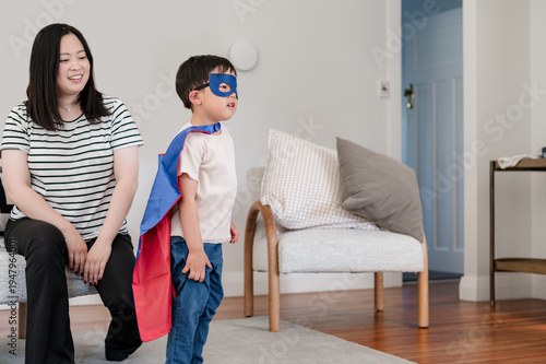Asian boy in superhero costume stands on a rug while a mum sits on a couch