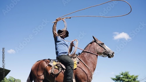 Cowboy sitting on a brown horse, expertly spinning a lasso over his head. Ranch hand showing roping skills on a sunny day with blue sky