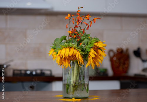 Bouquet of wilted sunflowers and orange flowers in glass jar on wooden table