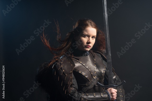A fierce warrior woman with fiery hair and a sword stands ready against a dark background, clad in detailed armor.