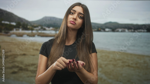 Woman with palms visible making pinched fingers and rubbing hand gesture on beach with coastal town in view; disappointment.
