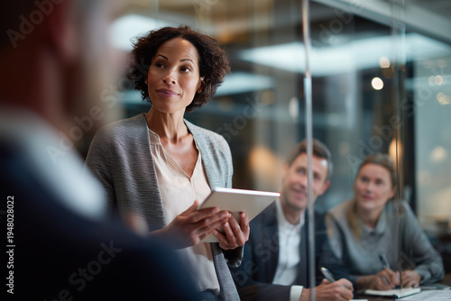 Team leader presenting ideas on digital tablet while coworkers listen and take notes in modern conference room.