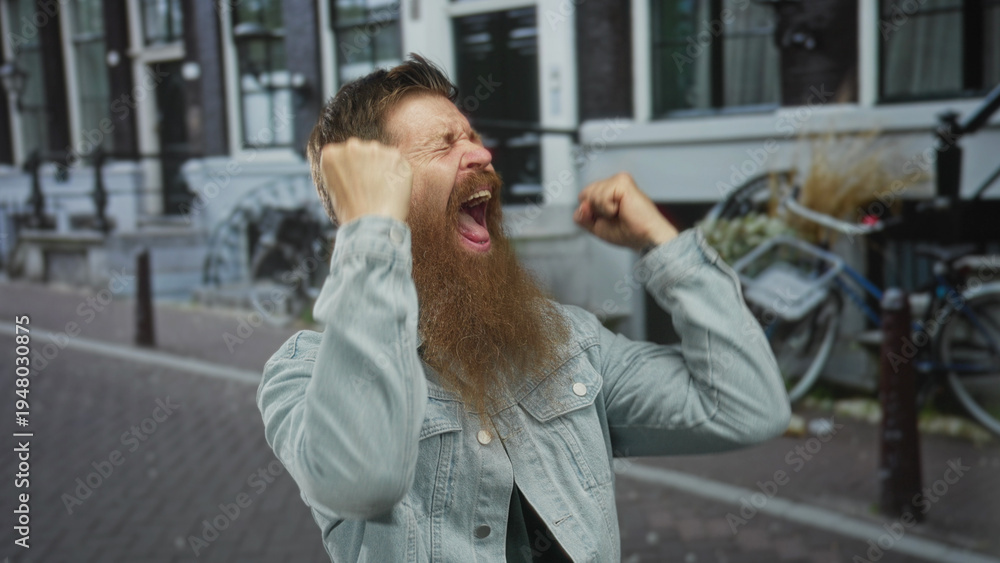 Fototapeta premium Bearded man in denim jacket clenches fists on a busy city street with brick buildings behind him; excitement.