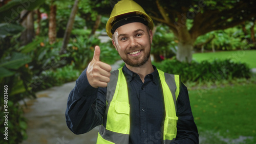 Man in hardhat and reflective vest giving thumbs up with hand near ear in forest; confidence teamwork.