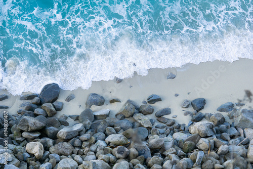 Фотография top view of white sea foam washing onto a sandy beach with large grey rocks scat