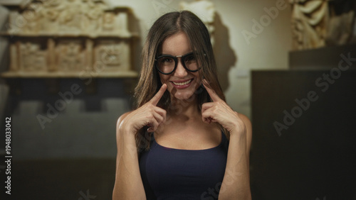 Fotografía Woman in blue tank top wearing glasses standing and smiling as she points finger to smile in building gallery; playful joy confidence warmth