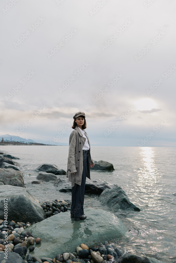 Fototapeta premium Young woman in casual outfit stands on rocky shore near calm sea under cloudy sky. Concept of solitude, nature, and peaceful outdoor lifestyle with soft natural light.
