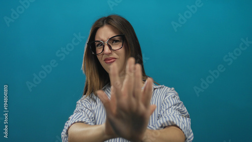 Obraz na płótnie Young hispanic woman in striped shirt wearing glasses pushes palm forward and frowns in blue studio; disgust
