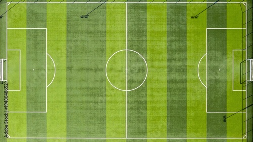 Image is a top-down view of a standard soccer field, featuring a symmetrical layout with clearly marked white lines on a vibrant green grass surface. The field is divided into two halves by a central 