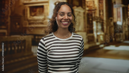 Free Download Woman smiling and tilting her head in an ornate church building, wearing a striped shirt and gold necklace with relaxed posture; joy faith warmth. - GFXLoad