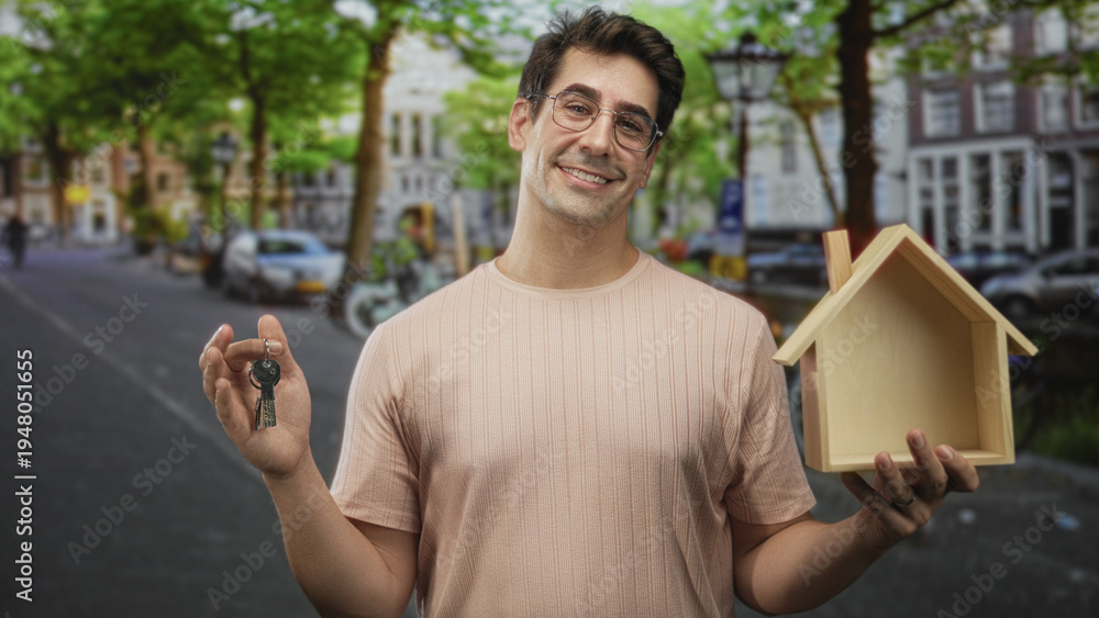 Fototapeta premium Man wearing glasses and a pale pink shirt holds a keyring and wooden house model, smiling and holding keys forward on a tree-lined city street with parked cars; homeownership confidence.