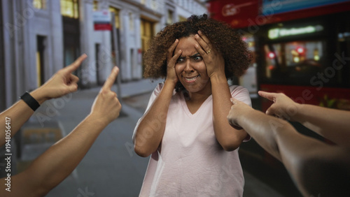 Woman holding her head with hands while surrounding hands point fingers at her on street near red bus; public shaming distress.