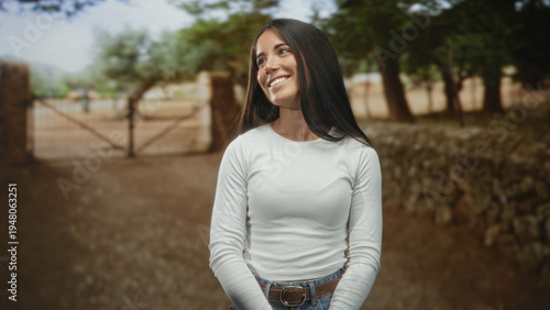 Photos Woman smiling, hands clasped, looking up beside a rustic gate on a street in white long sleeve top and jeans; serene contentment