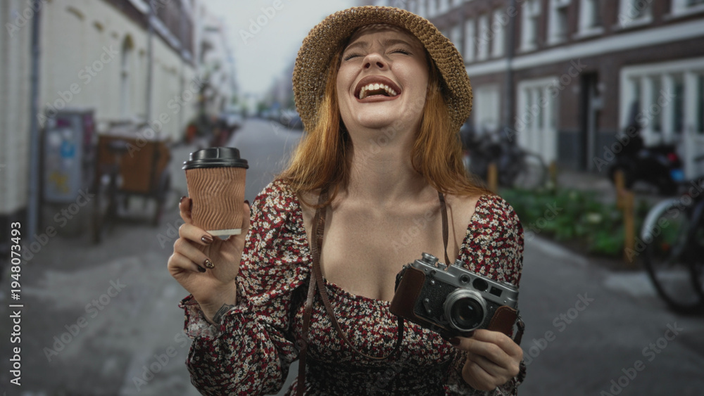 Fototapeta premium Redhead woman holding coffee cup and vintage camera, laughing with open mouth on cobblestone street, straw hat and floral dress visible; joyful travel moment.