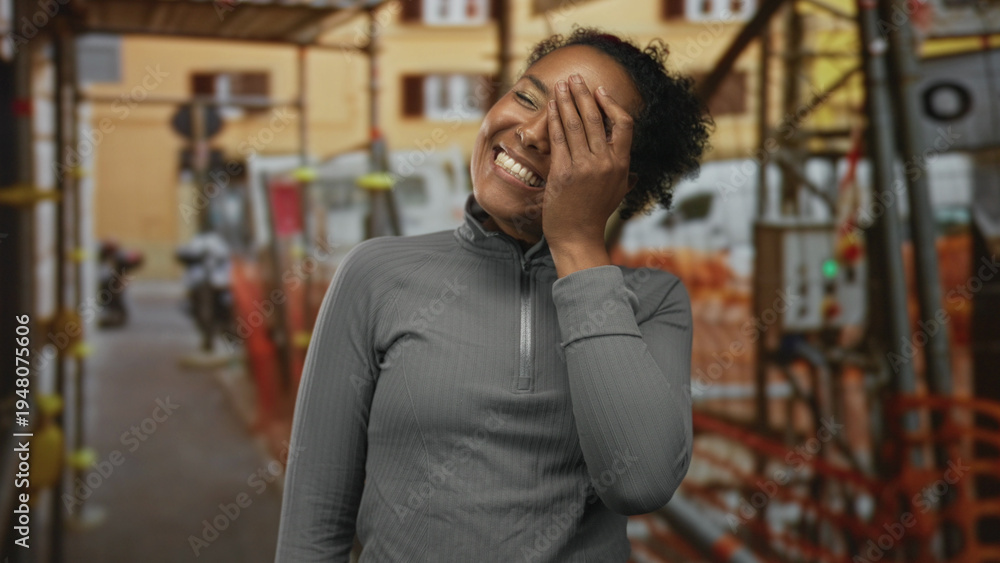 Fototapeta premium Young african american woman laughing and covering her face with hand at an outdoor building construction site scaffolding; joy.