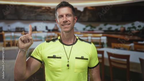Tableau sur toile Referee man wearing whistle and neon yellow shirt points index finger up in building restaurant interior, pocket detail visible, raised hand gesture; confidence authority