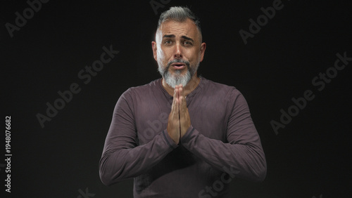 Photography Middle aged man with hands pressed together in a black studio, palms touching and pleading expression, bare hands visible; pleading apology