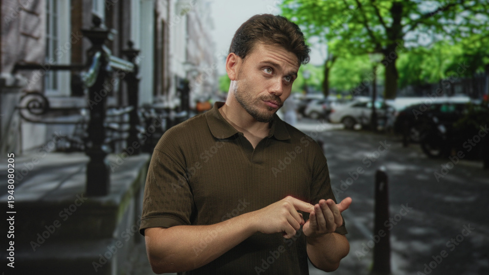 Fototapeta premium Young hispanic man wearing a brown polo pointing a finger to his palm on a leafy city street, smiling while tapping his open hand; casual happiness.