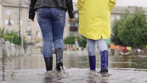 Photos girls in rubber boots are walking in a puddle, a flood on a city street, flood on the sides of roads and asphalt roadway, splashes different directions from wet muddy puddle, presence water districts
