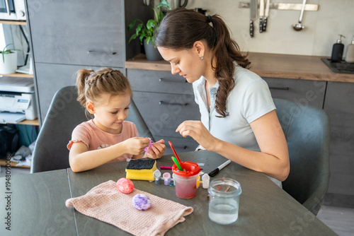 Mother and daughter decorating Easter eggs with colorful stickers while sitting at table at home. Family Easter craft activity, shared creativity and joyful holiday preparation indoors.