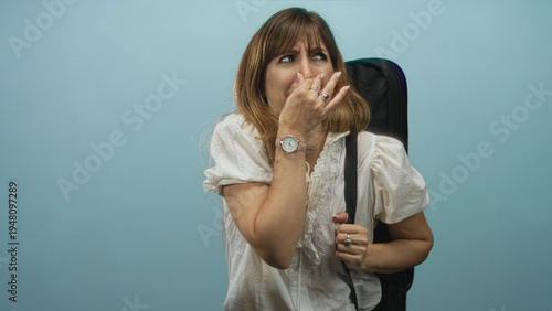 Foto Woman covering nose with hand while holding a case strap and wearing white ruffled blouse in studio with blue backdrop; odor smell disgust