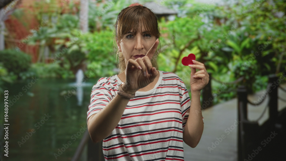 Fototapeta premium Woman points finger while holding a small red heart near chest on park walkway by green pond and bridge; love longing.