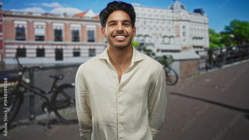 Fototapeta premium Man smiling and clasping hands beside parked bicycles on a street with historic buildings and a canal, facing camera and leaning slightly forward; joy gratitude welcome.