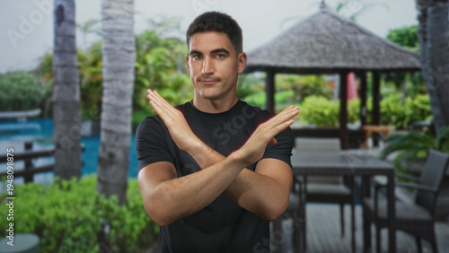 Принти на полотні Man with arms crossed x gesture at hotel pool gazebo wearing black tee and standing outdoors near palms and table; rejection