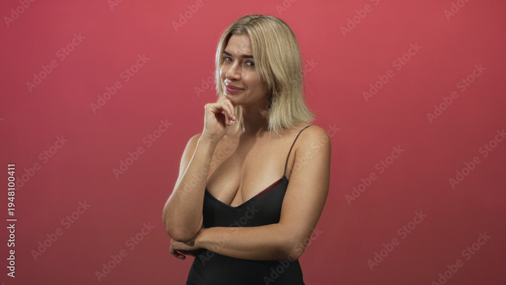 Fototapeta premium Young blonde woman touching chin with hand and crossed arms, wearing low-cut black tanktop against red studio backdrop; thoughtful contemplation.