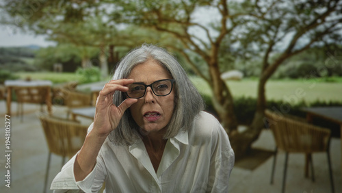 Foto Woman grey haired adjusting eyeglasses while squinting and leaning forward on a restaurant terrace outdoors; curiosity reflection